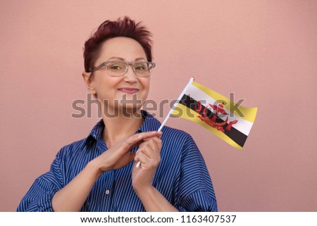 Brunei flag. Woman holding Brunei flag. Nice portrait of middle aged lady 40 50 years old with a national flag over pink wall background outdoors.
