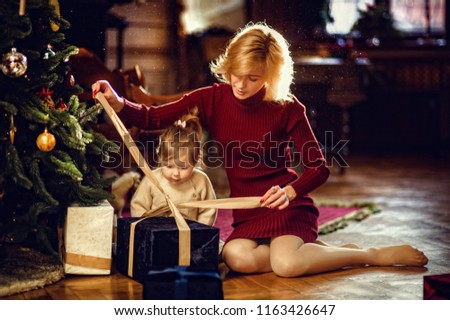 Blond hair mother in red dress and little toddler girl daughter untie a gift on new year morning sitting on floor