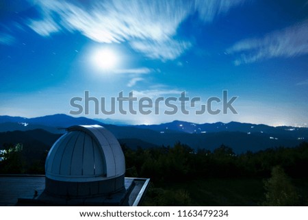 A panoramic view on an astronomical observatory in a cloudy night with a bright moond, with mountains landscape on background. 