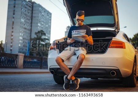 Distant plan of young man looking at map sitting in the truck of car.