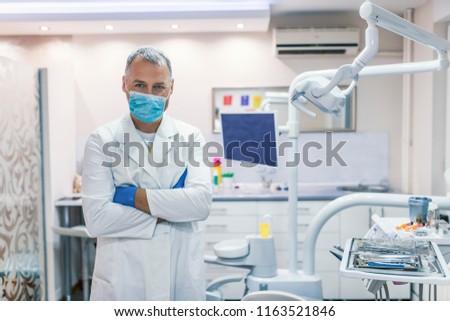 Male dentist standing with his hands crossed and looking at camera. Attractive male dentist in doctors white lab coat posing in modern dental office. Portrait of a friendly dentist  smiling
