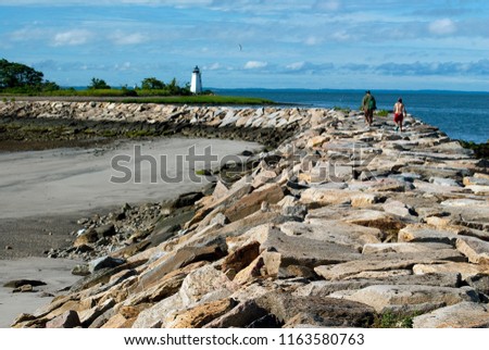 Young couple hiking along rocky breakwater towards Black Rock Harbor light connecting to Fayerweather Island in Bridgeport, Connecticut. It is a favorite challenge for many tourists to Seaside Park. Royalty-Free Stock Photo #1163580763