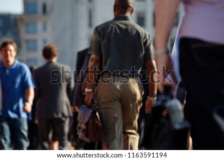 Black man in casual clothing walking to work carrying a leather satchel. Surrounded by other commuters. No point of image is totally in focus. London. Summer