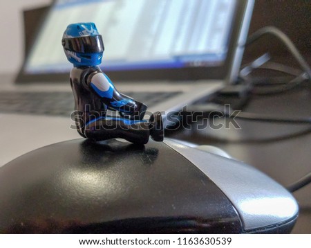 Plastic figure closeup riding a computer mouse with blurred  laptop and wires in the background ideal for fast computer, lets go signs technology learning IT study