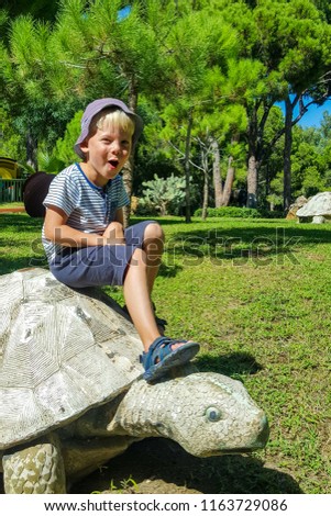 A laughing little boy sitting on a big turtle on a background of green grass, trees and blue sky.