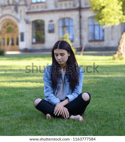 Brunette is sitting on the green lawn. Portrait of a cute girl in the park. Girl with long dark hair. 