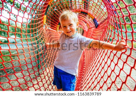 Blond scandinavian child having fun in red net tunnel in playground or amusement park. Holiday friendship and happy childhood concept.