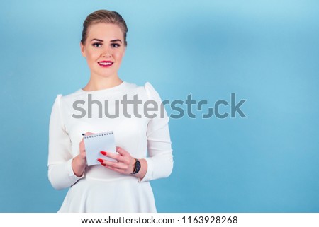 portrait of successful business woman blonde hairstyle perfect make-up red lips in stylish white suit writing in notebook notes planning in studio blue isolate