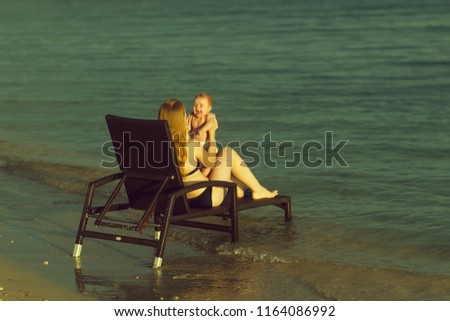 Two people back view of young blonde happy mother holding small cute smiling baby boy in hands playing together sitting on  chaise longue on sea water coast sunny weather outdoor, horizontal picture