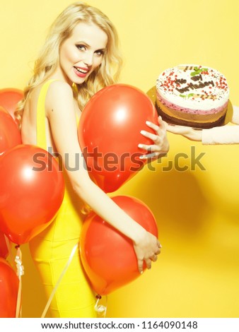 One attractive smiling young happy blond woman with long curly hair with birthday cake with candle in female hand near bunch of red party balloons in studio on yellow backdrop, vertical picture
