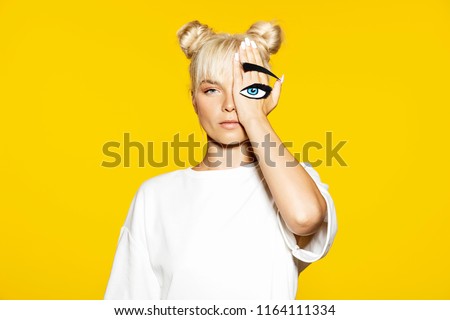 Portrait of serious caucasian female holding hand on face with fake eye and eyelash on it. Blonde model looking at camera in white shirt. Concept of emotions. Isolated on yellow background