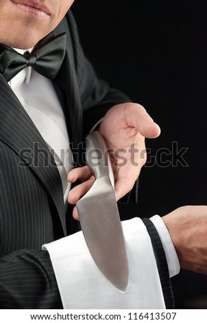 Close-up torso shot of a fine dining waiter in a bowtie and tux presenting a knife on the white pressed napkin over his arm.