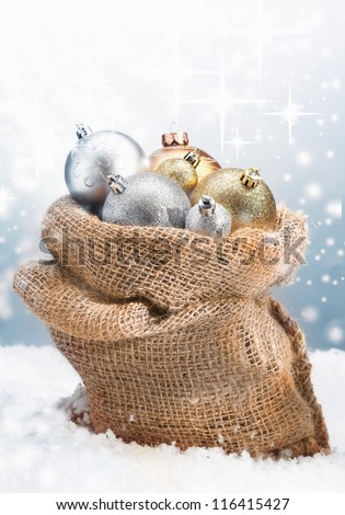 Winter Christmas baubles piled high in a rustic burlap bag nestled in fresh snow with a backdrop of twinkling stars on falling snowflakes