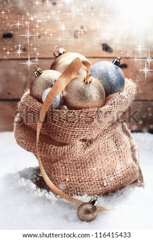 Decorative Christmas baubles piled high in a hessian bag on winter snow and decorated with a gold ribbon amidst dancing twinkling sunlight over a wooden wall