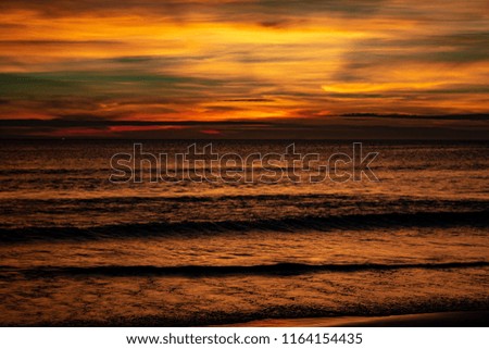 A tropical beach sunset the sun has passed the horizon .The clouds look painted with swirls of orange, blue, red and yellow. The ocean appears dark with highlights of colour. 