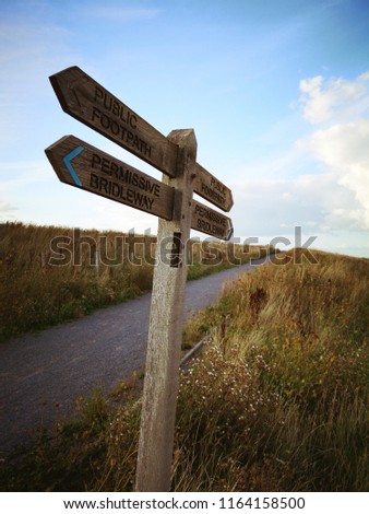 Permissive Bridleway and Public Footpath pointing wooden sign. Walking and hiking road, gravel path. Tall yellow green grass and blue sky with fluffy white clouds. Grey gravel bridleway. 