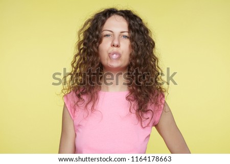 Playful funny American woman blows chewing gum, spends spare time with friends, wears short pink top, isolated over yellow background. Bored female with blue eyes inflates bubble gum in studio.