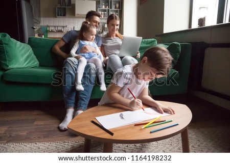 Creative kid boy son playing drawing with colored pencils while parents with little girl sister using laptop on sofa in living room, family children hobbies and activities at home on weekend concept