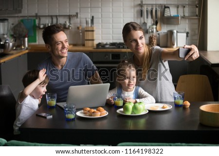 Happy family with children taking selfie on phone having breakfast with devices together in the kitchen, smiling mom dad and kids posing waving hands making photo self-portrait on smartphone at home