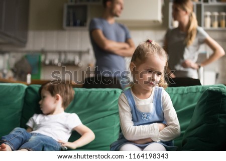 Upset little girl feeling sad after fight with brother sitting on sofa with worried parents on background, sulky frustrated sister ignoring child boy disinterested or bored, siblings rivalry concept Royalty-Free Stock Photo #1164198343