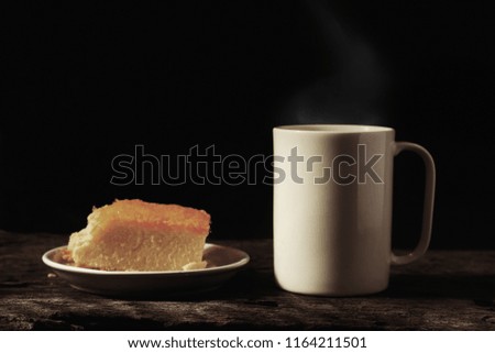 A cup of white coffee with steam from the heat and a slice of cake in a dish placed on an old wooden floor. On a black background