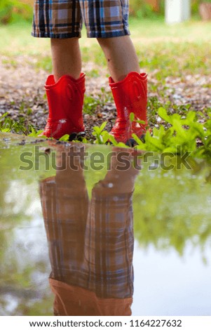 Legs of child wearing pair of red rubber boots with reflection in water puddle