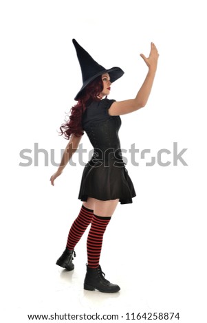 full length portrait of red haired girl wearing black witch costume and pointy hat.  standing pose, with back to the camera. isolated on white studio background.