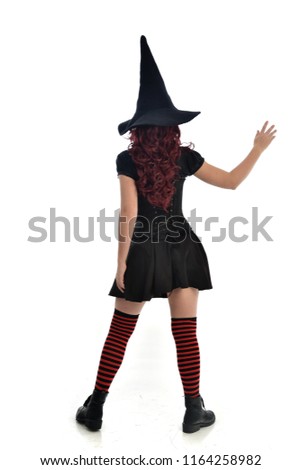 full length portrait of red haired girl wearing black witch costume and pointy hat.  standing pose, with back to the camera. isolated on white studio background.