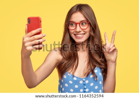 Beautiful female youngster makes peace gesture while poses for making selfie, uses modern cell phone, has toothy smile, wears red spectacles and blue polka dot blouse, poses over yellow background