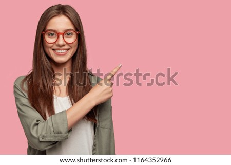 Satisfied young female with positive expression, attracts your attention at copy space aside, demonstrates some items in shop, dressed in casual shirt, has dark straight hair, isolated over pink wall Royalty-Free Stock Photo #1164352966