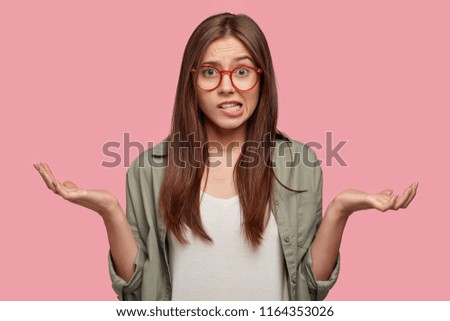 Indifferent puzzled female shruggs shoulders, purses lips, has clueless expression, cant make decision, chooses between two items, feels confused, dressed in casual clothes, isolated on pink wall Royalty-Free Stock Photo #1164353026