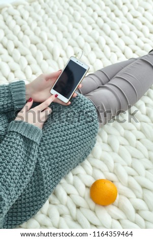 Very cozy picture for the autumn, September, October, November. A girl in a warm green knitted sweater dials SMS, uses mobile Internet, plays on the phone. An orange on a blanket.