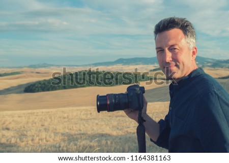 Man professional photographer with digital camera walking in tuscany fields, taking photos. Photo Camera in male hands over summer landscape in Tuscany with fields, blue sky,moutains, Italy, Europe
