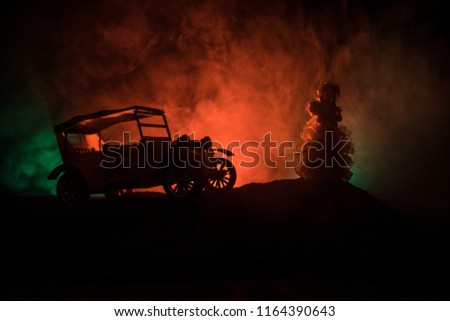 Silhouette of old vintage car and old style weared woman in dark foggy toned background with glowing lights in low light. Selective focus