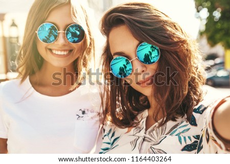 Two young female stylish hippie brunette and blond women models in summer hipster clothes taking selfie photos for social media on smartphone on the street background