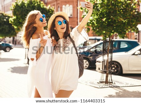 Two young female smiling hippie brunette and blond women models in summer sunny day in white hipster clothes taking selfie photos for social media on smartphone on the street background. 
