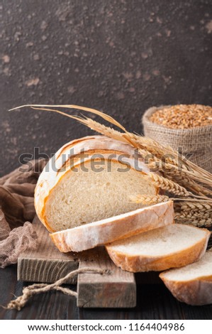 Loaf of white wheat bread, sliced on a cutting board on a brown wooden table with a bag of wheat grains and wheat spikelets, spikes. Copy space. Front view. Close up. Home bakery. Homemade. 