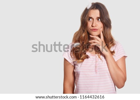 Horizontal shot of thoughtful woman has troubles expression, being deep in thoughts, has feathers on head, dressed in nightclothes, stands against white background with copy space for your text