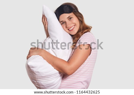 Positive European woman with friendly smile leans at white pillow, wears sleep mask and pyjamas, rejoices having good rest at night, stands sideways against white wall. Good morning concept.
