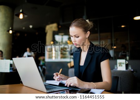 Serious woman economist writing notary in notepad during work on laptop computer while sitting in restaurant during work break. Female employee writing a monthly report in notebook from net-book 