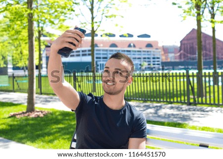 Handsome male making selfie photo on mobile phone camera while sitting in city park in sunny summer day. Smiling man having online video call on smartphone during free time outdoors 