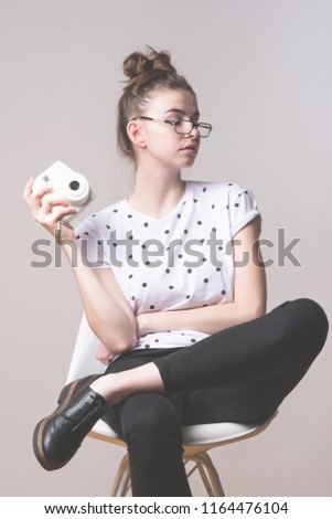 Cool millennial young hipster teenage girl photographer sitting on chair, wearing glasses, holding instant photo camera. Matte filter, closeup, studio lighting, no retouch.