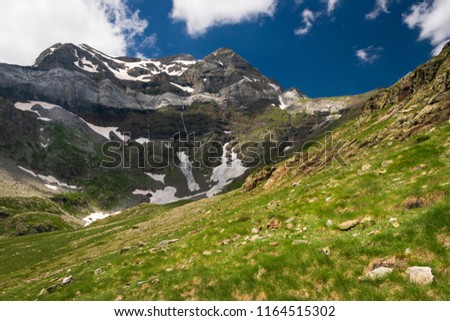 View of a mountain range in Spanish Pyrenees, with green meadows in the foreground and deep blue sky. The photo was taken in summer, on the trail from the village Parzán to the Cirque de Barroude.