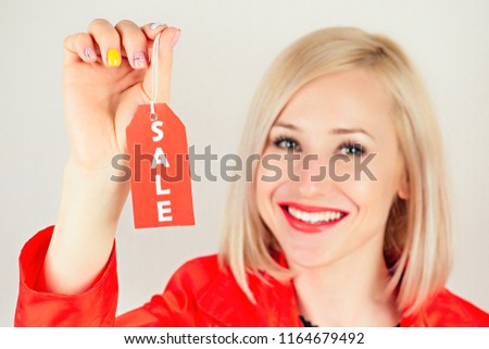 beautiful smiling blonde woman shopaholic holding a red label ( tag , tab , tally ) with sale inscription close-up in her hand . concept of seasonal sales and shopping