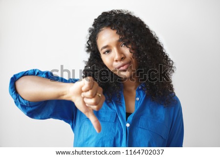 Negative human reaction, attitude and life perception. Serious displeased young dark skinned female with loose curly hair making thumbs down gesture, demonstrating her dislike and disrespect