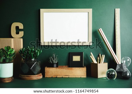 Green modern and stylish room with wooden mock up poster frame, office accesories, boxes and plants. Creative composition of desk in green interior.