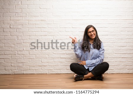 Young indian woman sit against a brick wall pointing to the side, smiling surprised presenting something, natural and casual