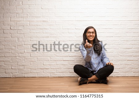 Young indian woman sit against a brick wall inviting to come, confident and smiling making a gesture with hand, being positive and friendly