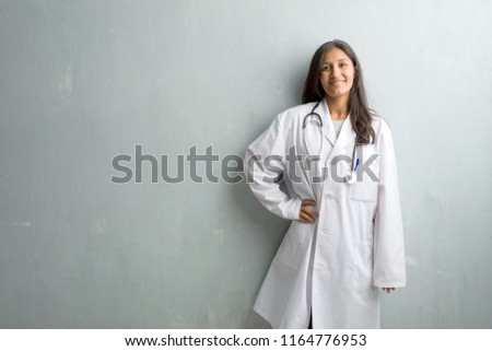 Young indian doctor woman against a wall with hands on hips, standing, relaxed and smiling, very positive and cheerful