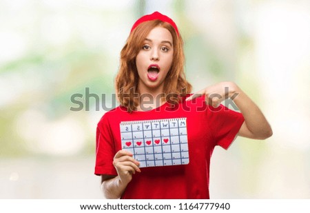 Young beautiful woman over isolated background holding menstruation calendar with surprise face pointing finger to himself
